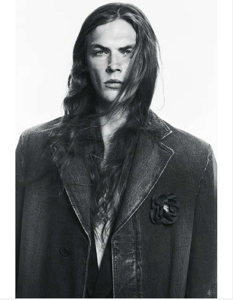 Monochrome portrait of a man with long wavy hair wearing a denim jacket with a flower brooch against a white background.