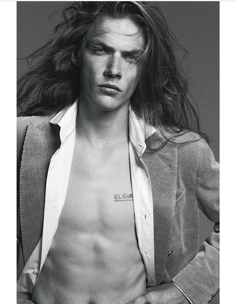 Black and white portrait of a shirtless man with long hair wearing a corduroy jacket, posing indoors against a neutral backdrop.