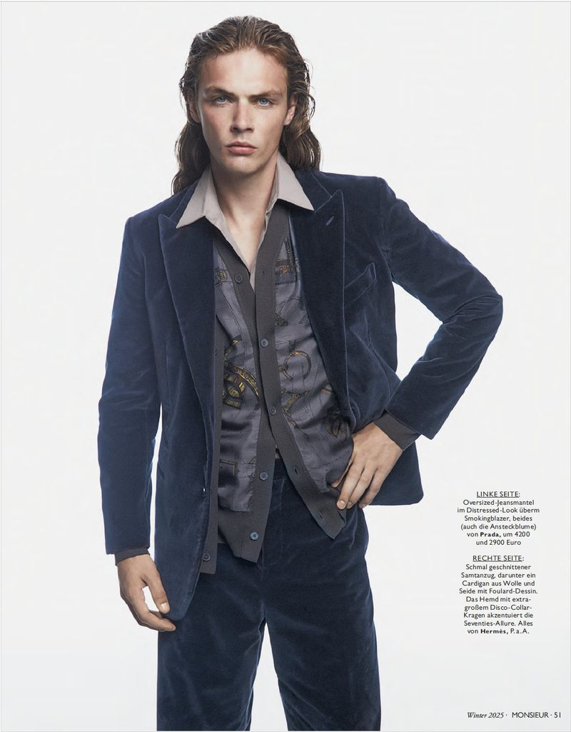 Editorial fashion shot of a young adult male model in a velvet suit, unbuttoned shirt, and long brown hair against a white background.