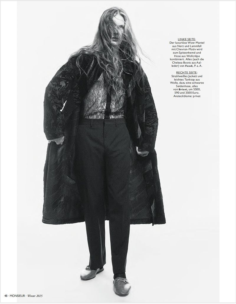 Fashion model with long hair wearing a dark patterned overcoat, sheer lace shirt, and dark trousers against a white studio background.