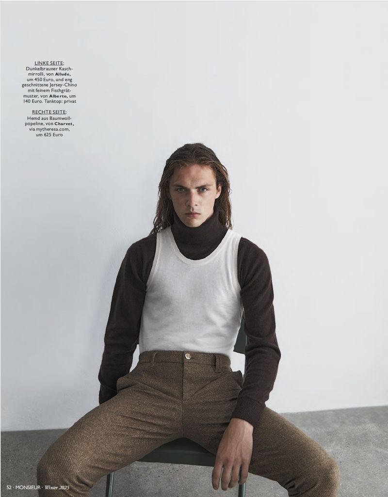 A man with shoulder-length hair sits on a chair wearing a white tank top over a dark turtleneck and brown trousers in a studio.
