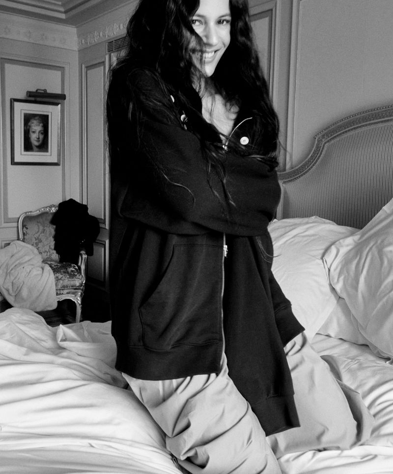 A person with long dark hair, wearing a zip-up sweatshirt and pants, smiles while standing on a bed in a room with classic decor.