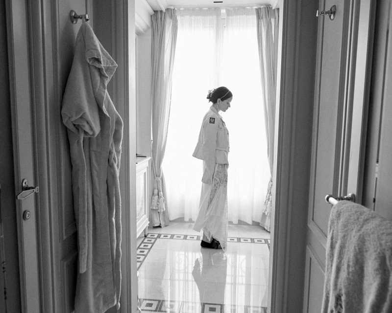 A woman in a white robe stands in a bathroom near a window, with a bathrobe hanging on a door, in a monochrome photograph.