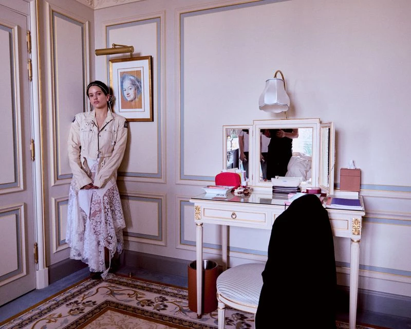 A woman in a cream jacket and lace skirt stands in a corner of a room with a vanity, framed artwork, and patterned carpet.