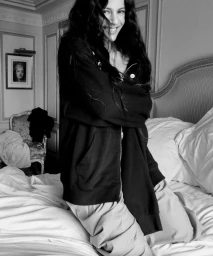 Rosalía smiling while kneeling on a white bed wearing a dark hooded zip-up sweatshirt in an elegant hotel room interior