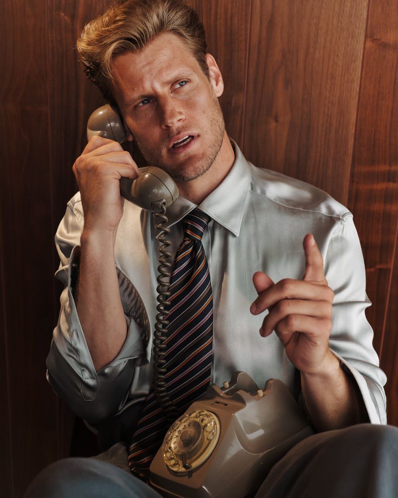 Man in shirt and tie using rotary phone indoors, vintage style, against wood paneling, with serious expression and raised finger.