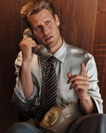 Man in shirt and tie using rotary phone indoors, vintage style, against wood paneling, with serious expression and raised finger.