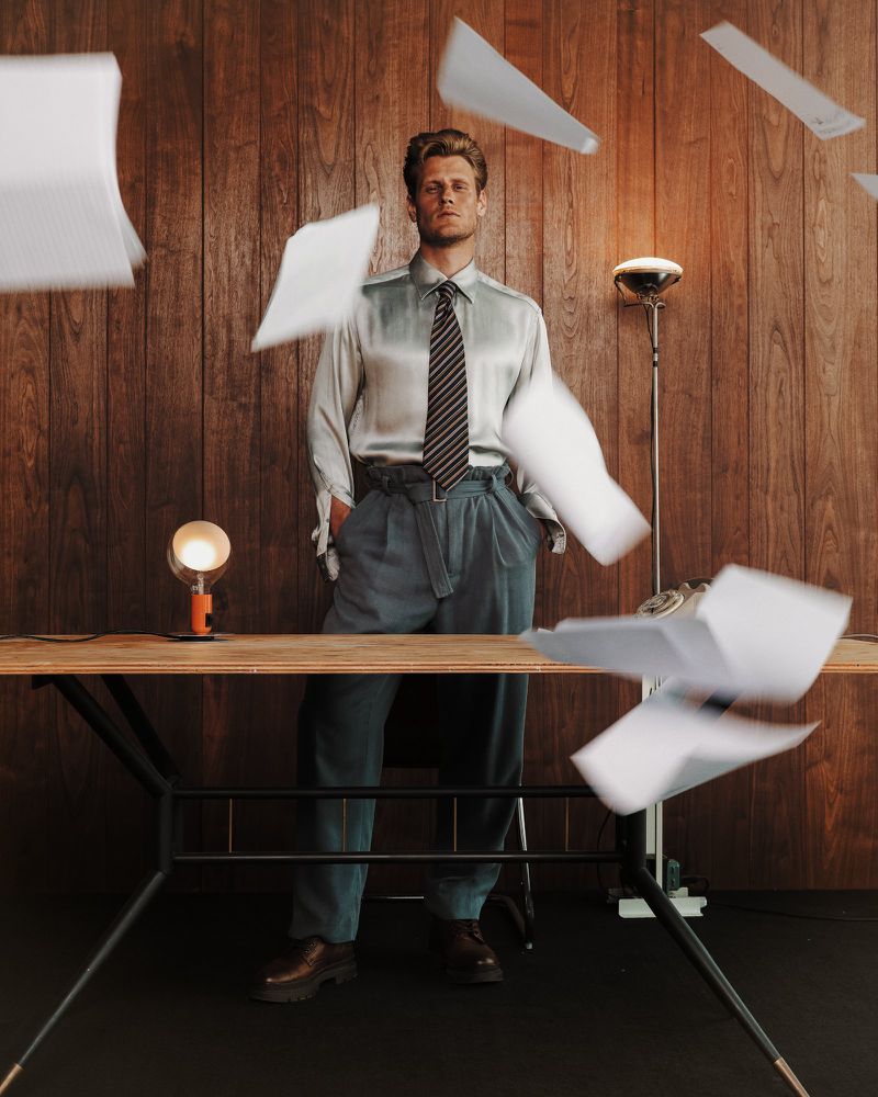 Adult male standing at a desk throwing papers into the air in front of a wood-paneled wall in an indoor setting.