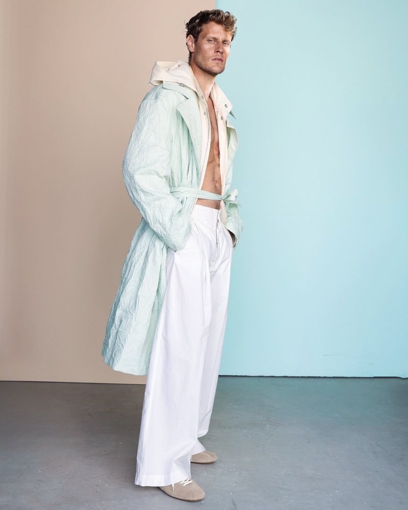 Full body shot of an adult male model wearing a light blue trench coat and white pants in front of a split color background.