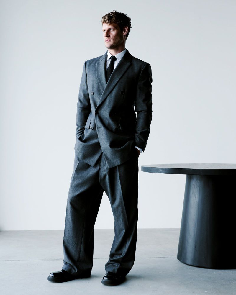 Portrait of an adult man in a grey double-breasted suit with a black tie, standing in front of a white wall with a black table.