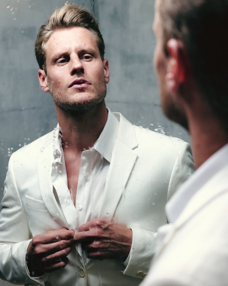 A blonde adult male fastens the buttons of a white shirt and jacket in front of a mirror, with a blurred grey background.