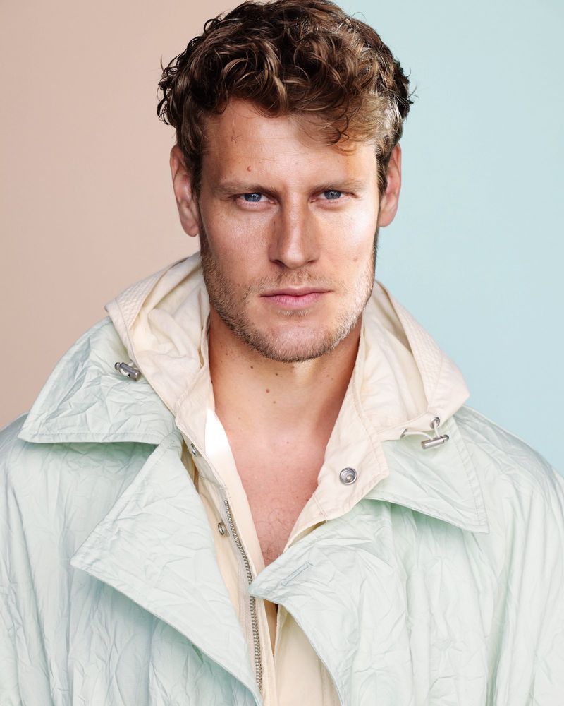 Portrait of an adult male with wavy brown hair wearing a pale green and cream jacket, posing against a split peach and turquoise background.