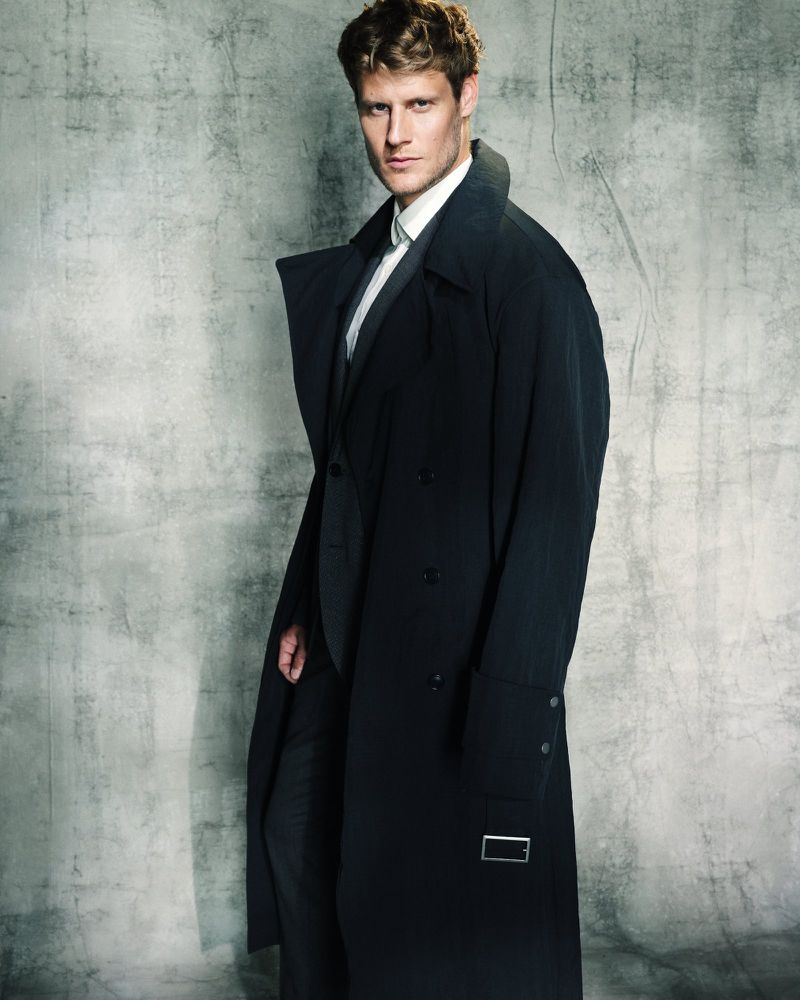A stylish, blonde-haired man in a suit and black trench coat poses in front of a textured grey background. Fashion photography.