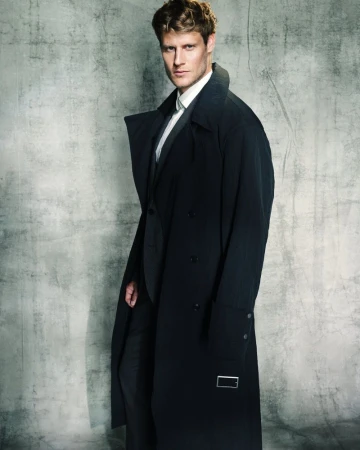 A stylish, blonde-haired man in a suit and black trench coat poses in front of a textured grey background. Fashion photography.