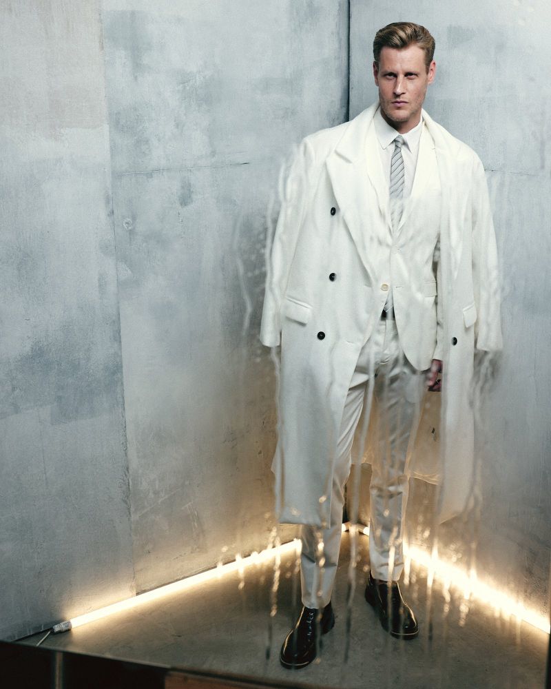 Adult male poses indoors wearing a white three-piece suit and overcoat, standing near a gray concrete wall with concealed lighting.