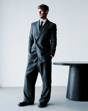 Man in grey double-breasted suit and black tie standing next to a round dark table against a white background