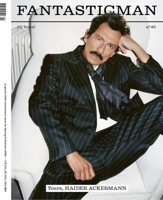 Man with mustache in pinstripe suit poses on white chair for magazine cover, looking at camera in studio with dramatic lighting.