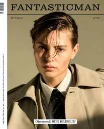 Close-up portrait of model Bibi Breslin wearing a beige trench coat, collared shirt, and dark tie against an olive background.