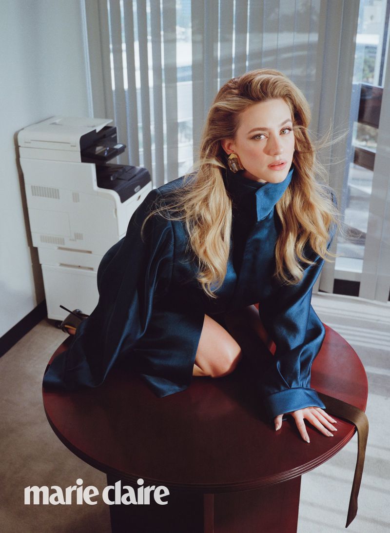 Portrait of a blonde woman in a blue jacket sitting on a table in an office with a printer in the background, photographed for Marie Claire.