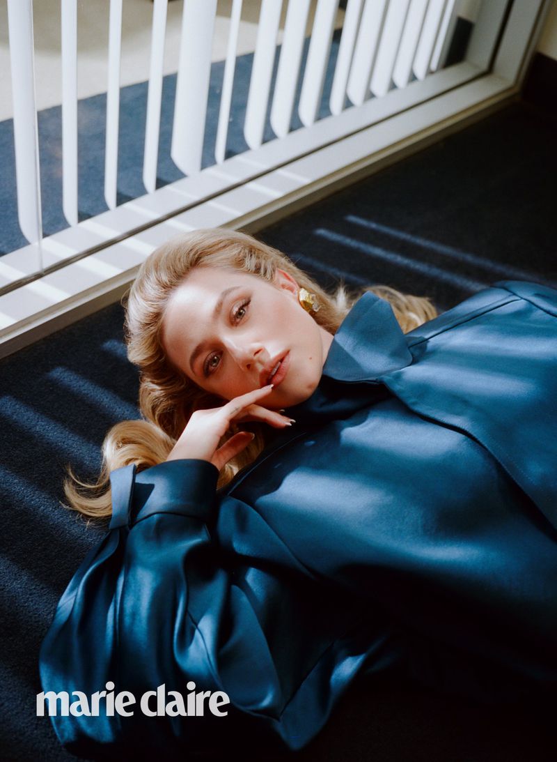 Indoor fashion shot of a blonde woman in a blue top lying on a dark blue surface by a white slated partition with soft studio lighting.