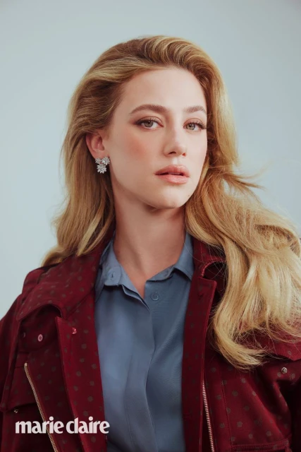 Portrait of a blonde woman in a jacket and blouse, featuring blonde hair, light makeup, and jewelry, set against a light backdrop.