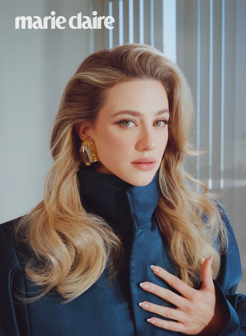 Portrait of Lili Reinhart wearing a dark blue high collar coat and gold earrings with blonde voluminous hair
