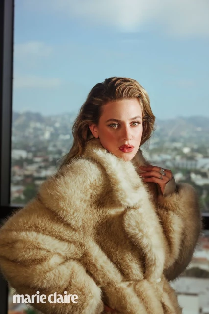 Portrait of Lili Reinhart wearing a fur coat, against a background of city buildings and blue sky, with the 'marie claire' logo in the corner.