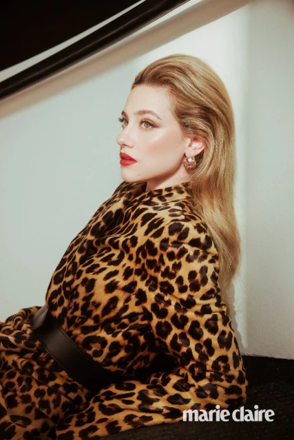 Portrait of a blonde woman in a leopard-print jacket, black belt, and red lipstick, looking off to the side in an indoor studio setting.