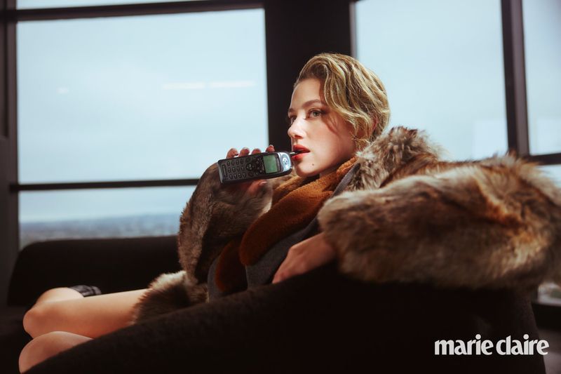A blonde-haired woman with red lipstick, wearing a fur coat, reclines on a couch with a vintage phone, set against a blurred city backdrop.