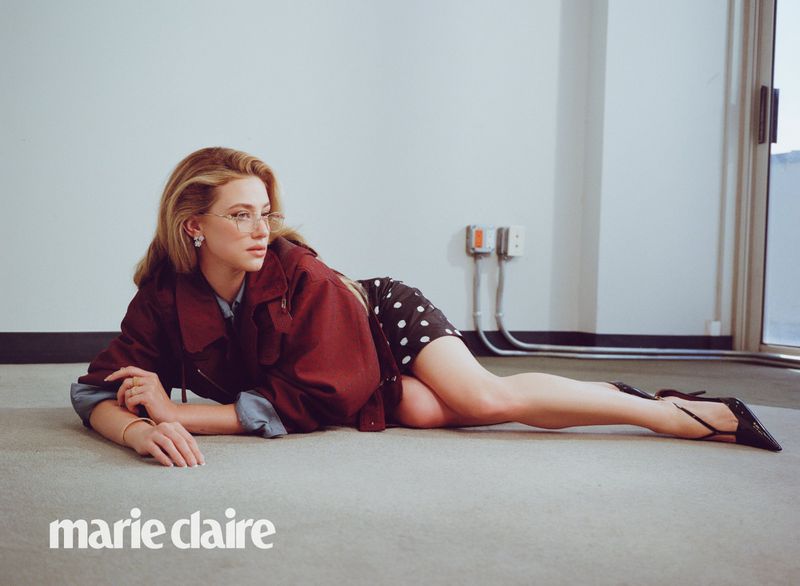 A woman in glasses and a polka-dot skirt lies on the floor in a maroon jacket with a magazine logo visible in the lower left.