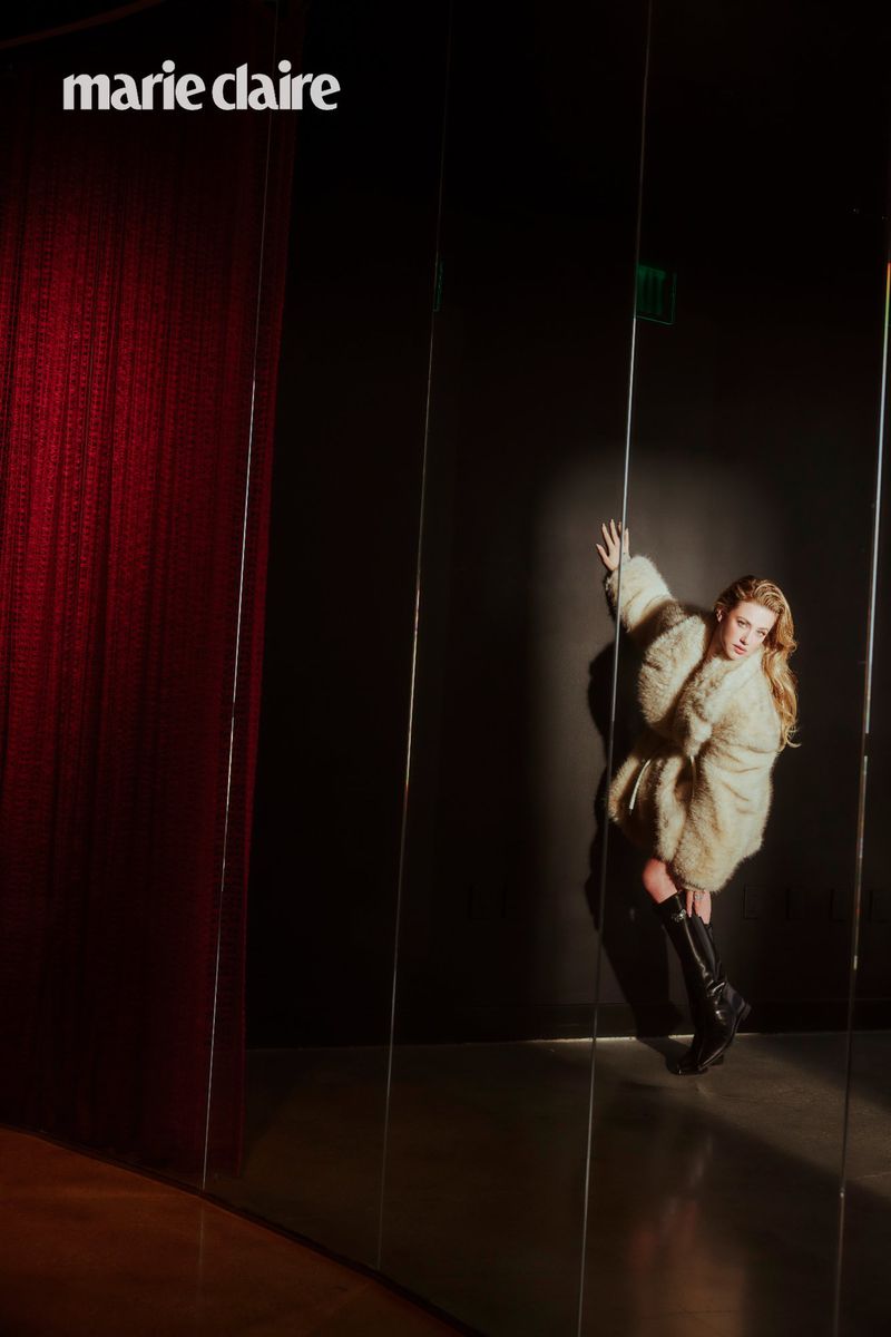Fashion editorial shot of a blond woman wearing a fur coat and black boots, posing in front of a mirrored wall and red curtain.