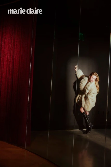 Fashion editorial shot of a blond woman wearing a fur coat and black boots, posing in front of a mirrored wall and red curtain.