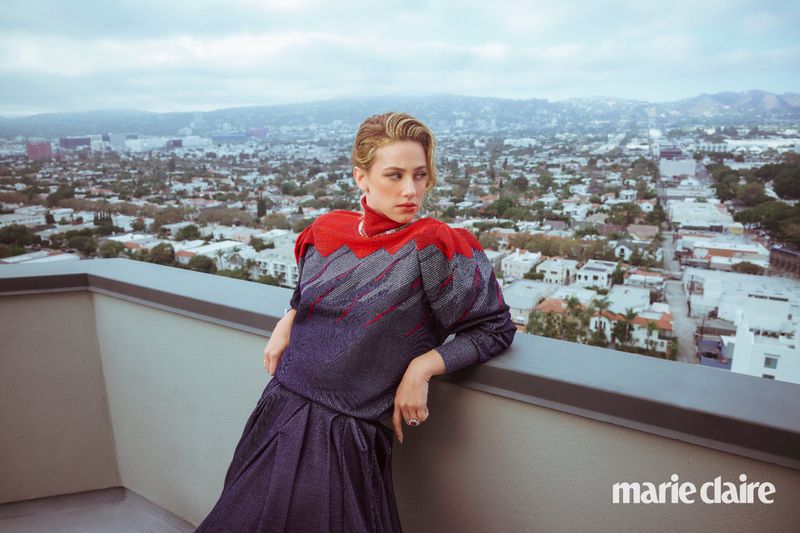 Blonde woman in a red and gray dress poses on a balcony overlooking a city, wearing jewelry and light makeup.