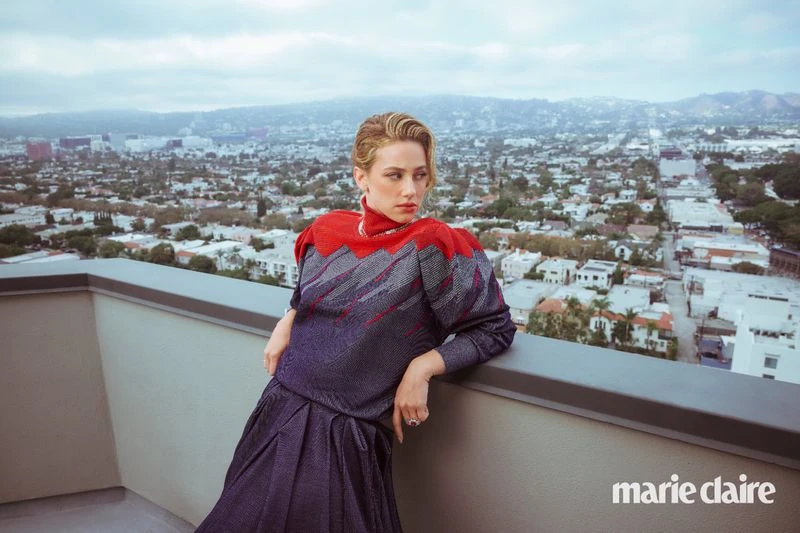 Blonde woman in a red and gray dress poses on a balcony overlooking a city, wearing jewelry and light makeup.