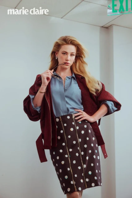Adult female poses indoors wearing a button-down shirt, burgundy jacket, and polka dot skirt, holding glasses in her mouth.