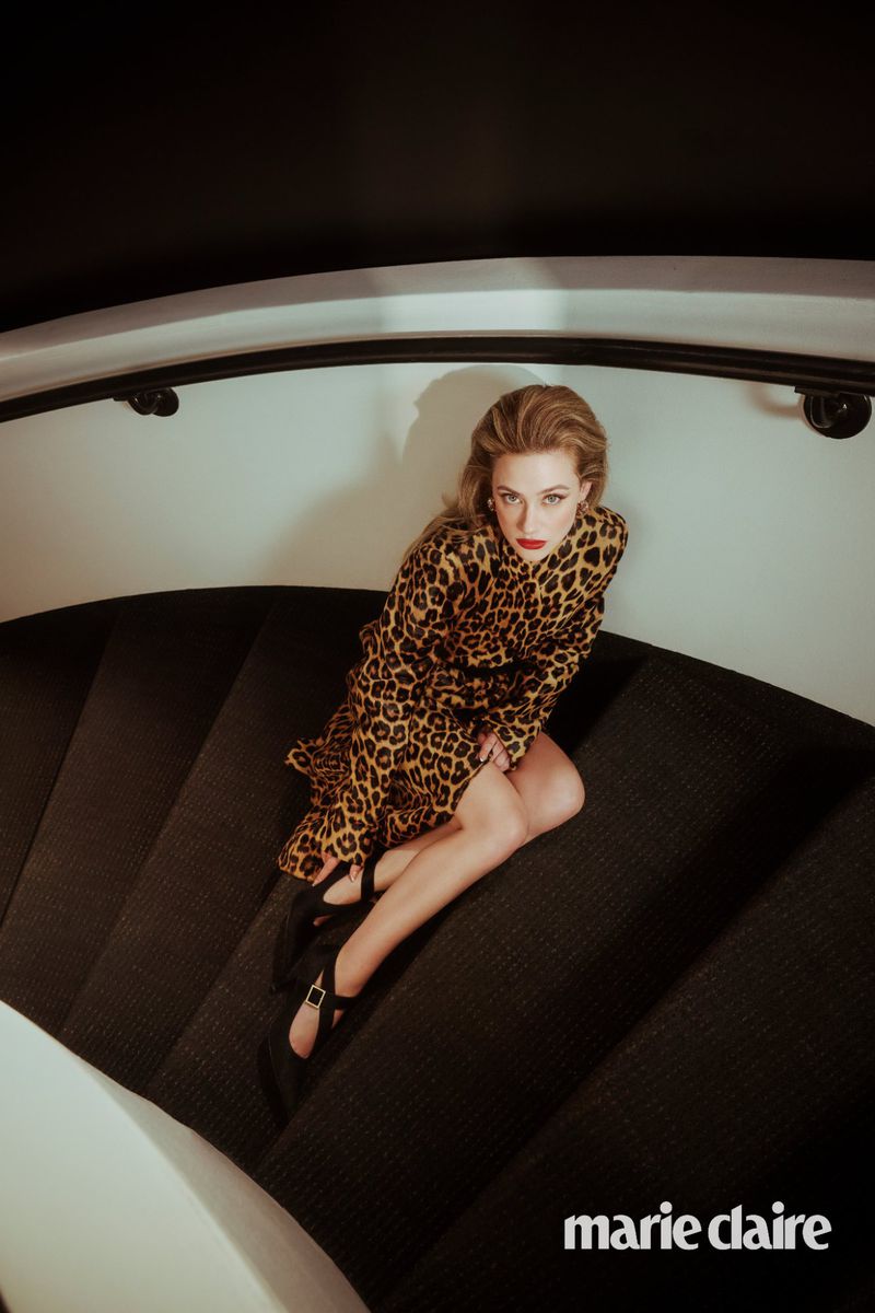 Blonde woman in leopard print dress sits on carpeted stairs, wearing Mary Jane shoes and red lipstick in a beauty fashion editorial.