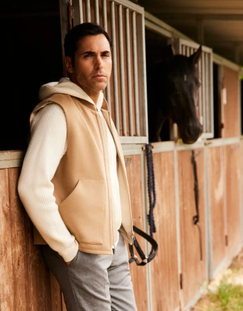 Adult man in a beige vest and gray pants leans against a wood horse stall; a dark horse stands inside the stall, rural setting.