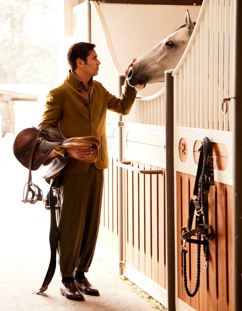 Equestrian lifestyle portrait of a man petting a white horse inside a stable, holding riding gear, dressed in tan jacket and olive pants.