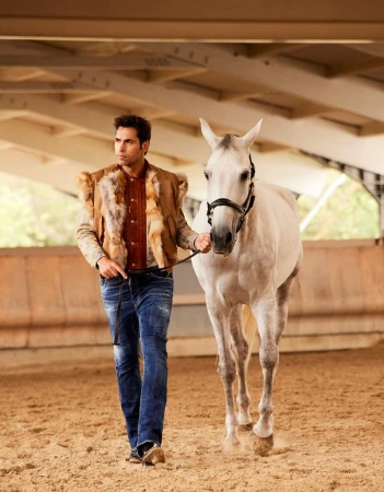 A man in a fur vest walks a white horse in an indoor arena, showcasing the equestrian style and the bond between human and animal.