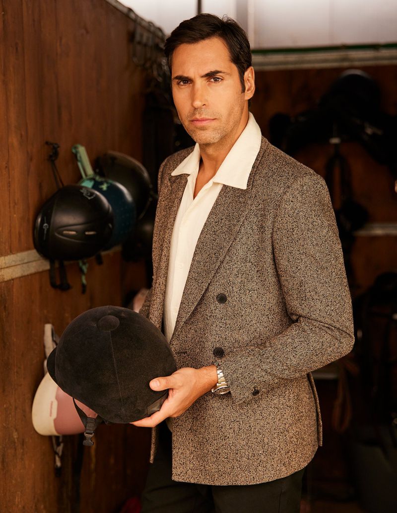 Portrait of an adult male in a tweed blazer holding a riding helmet, standing in front of a wood-paneled wall with other helmets hanging.