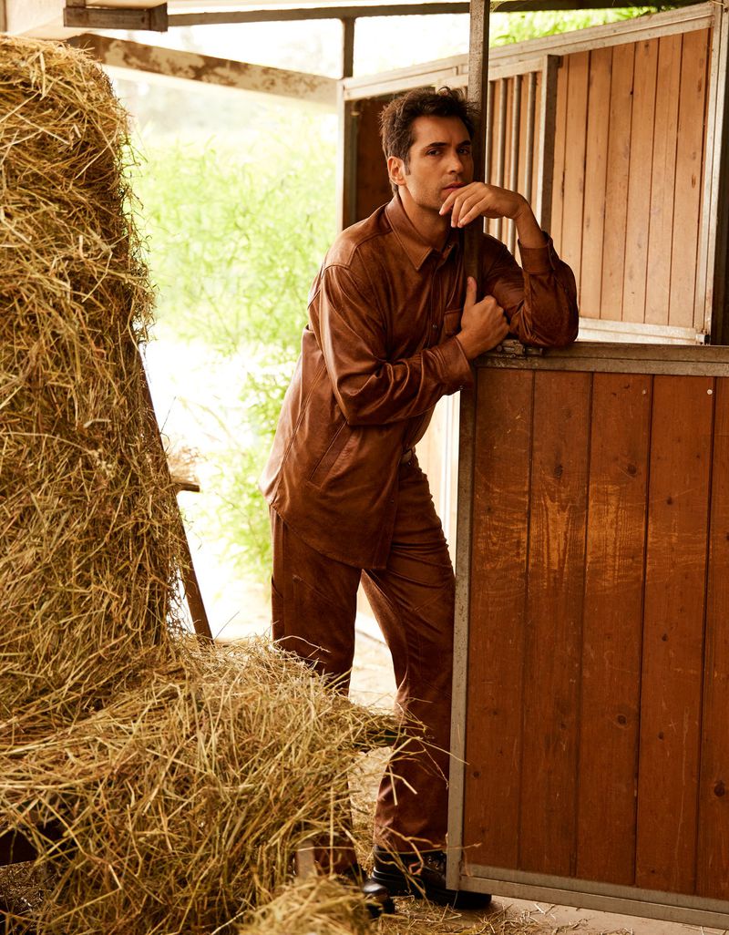 Male model in brown jumpsuit leaning on wood structure in a barn with hay bales, gazing into the distance with a contemplative expression.