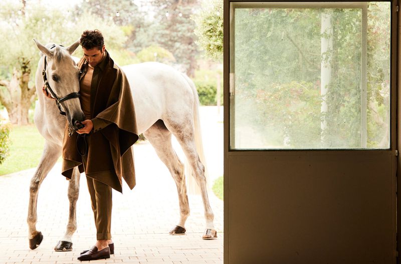 White horse with a man wearing a brown poncho and matching pants standing in a doorway with green trees in the background.