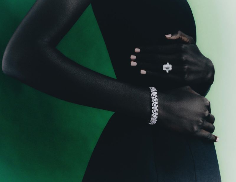 Elegant dark-skinned model showcasing a sparkling diamond ring and floral bracelet against a gradient green backdrop in a sophisticated fashion shot.