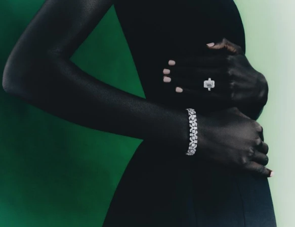 Elegant dark-skinned model showcasing a sparkling diamond ring and floral bracelet against a gradient green backdrop in a sophisticated fashion shot.