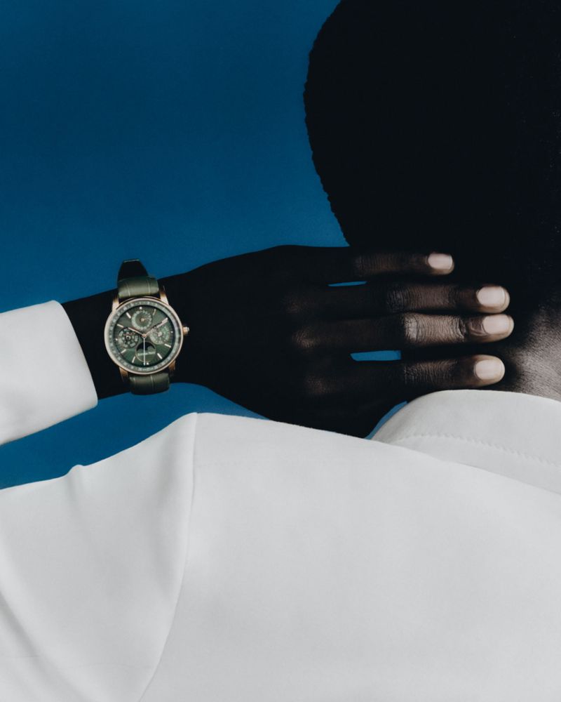 Close-up studio shot of a dark-skinned adult with their hand behind their head, wearing a watch and a white long sleeve shirt.