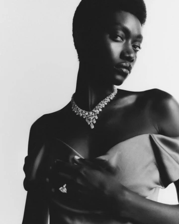 Striking portrait of a Black woman in monochrome, wearing a jeweled necklace and ring, standing against a bright, neutral background.