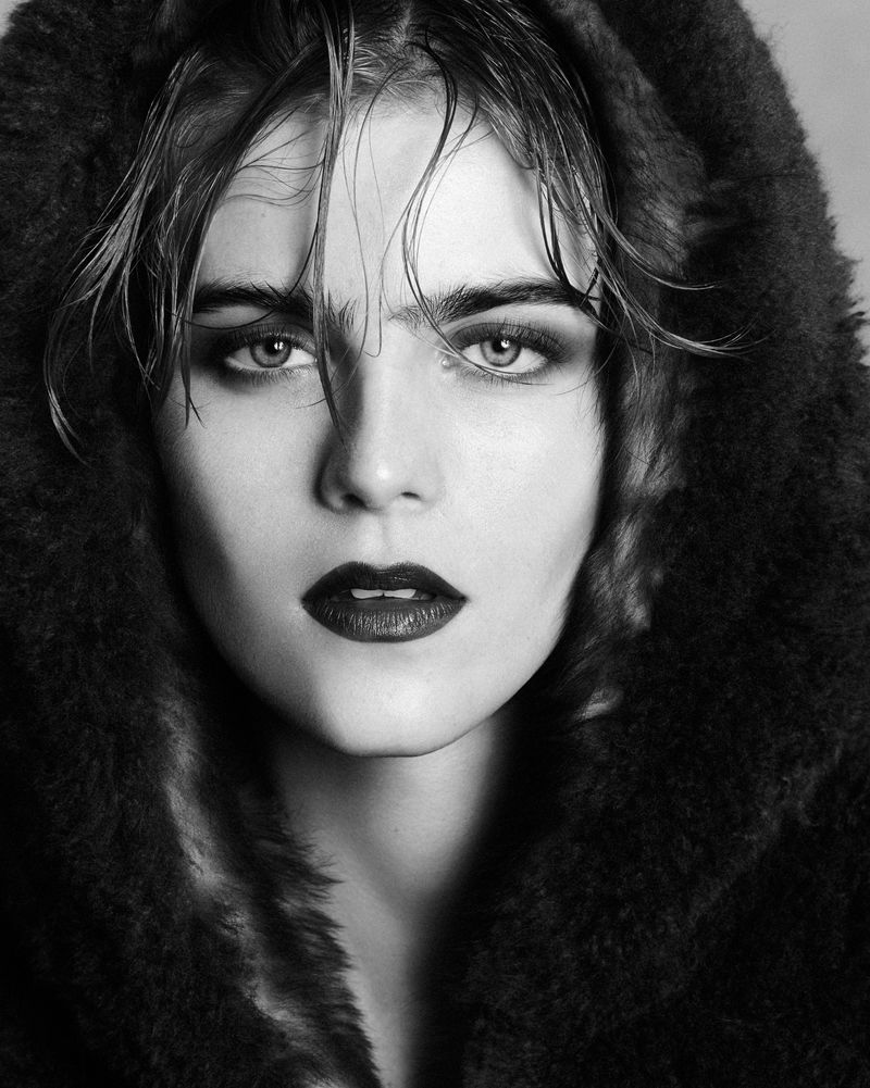 Striking black and white portrait of a woman with dark lipstick wearing a furry hooded jacket, capturing a high fashion beauty look.
