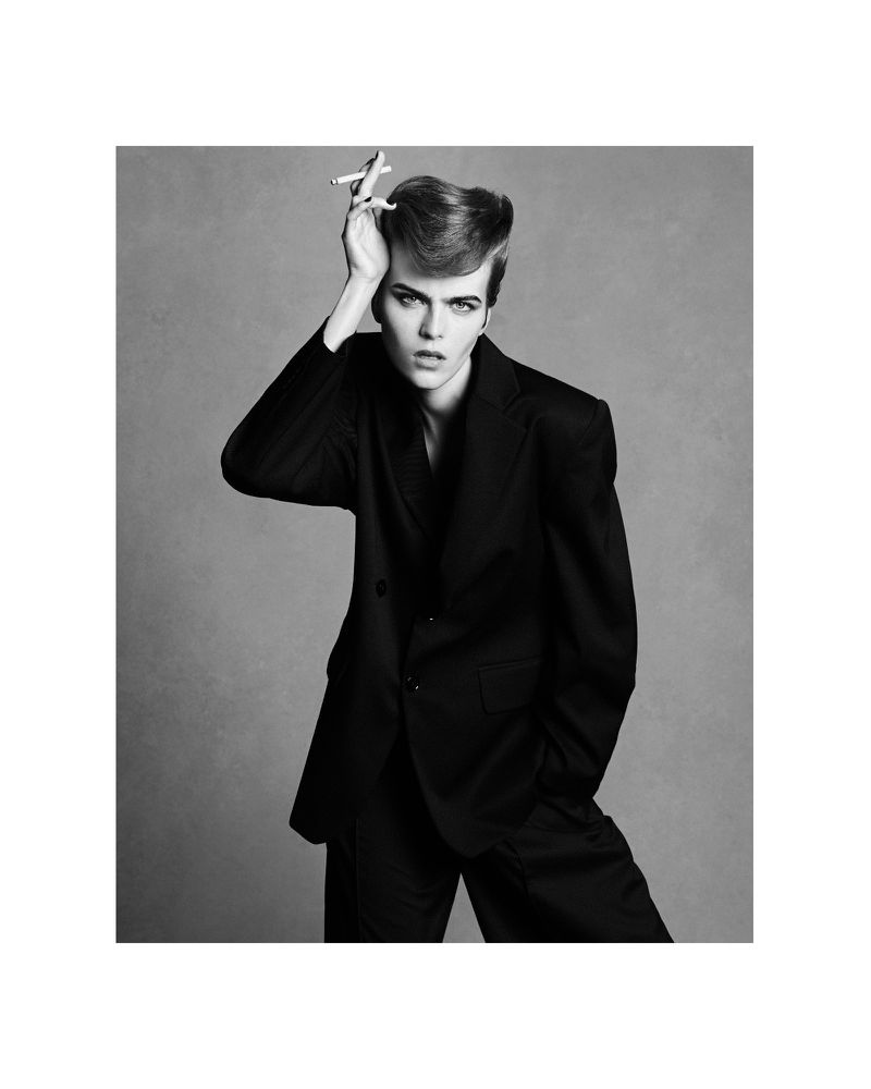 Black and white portrait of a young adult with an elaborate pompadour wearing a dark suit and holding a cigarette to his head.