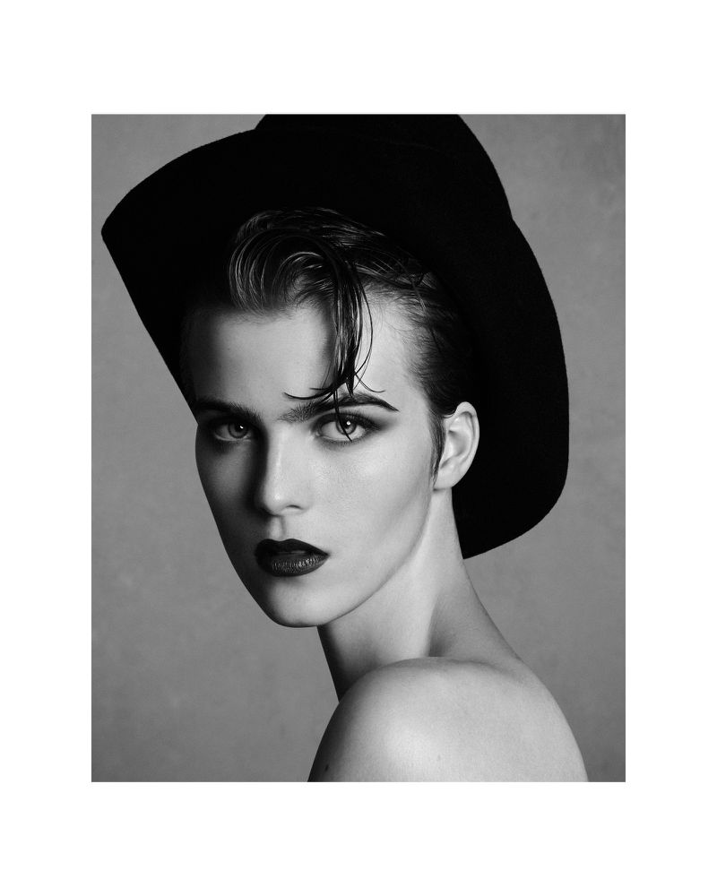 Monochrome portrait of a young adult woman with fair skin wearing a black hat, dark lipstick, and heavy eye makeup against a grey background.