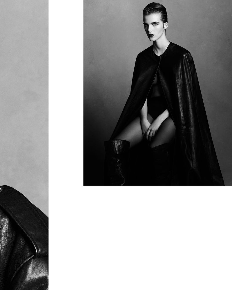 Monochrome studio portrait of a female model wearing a black bodysuit, thigh-high boots, and a black leather cape, striking a pose.